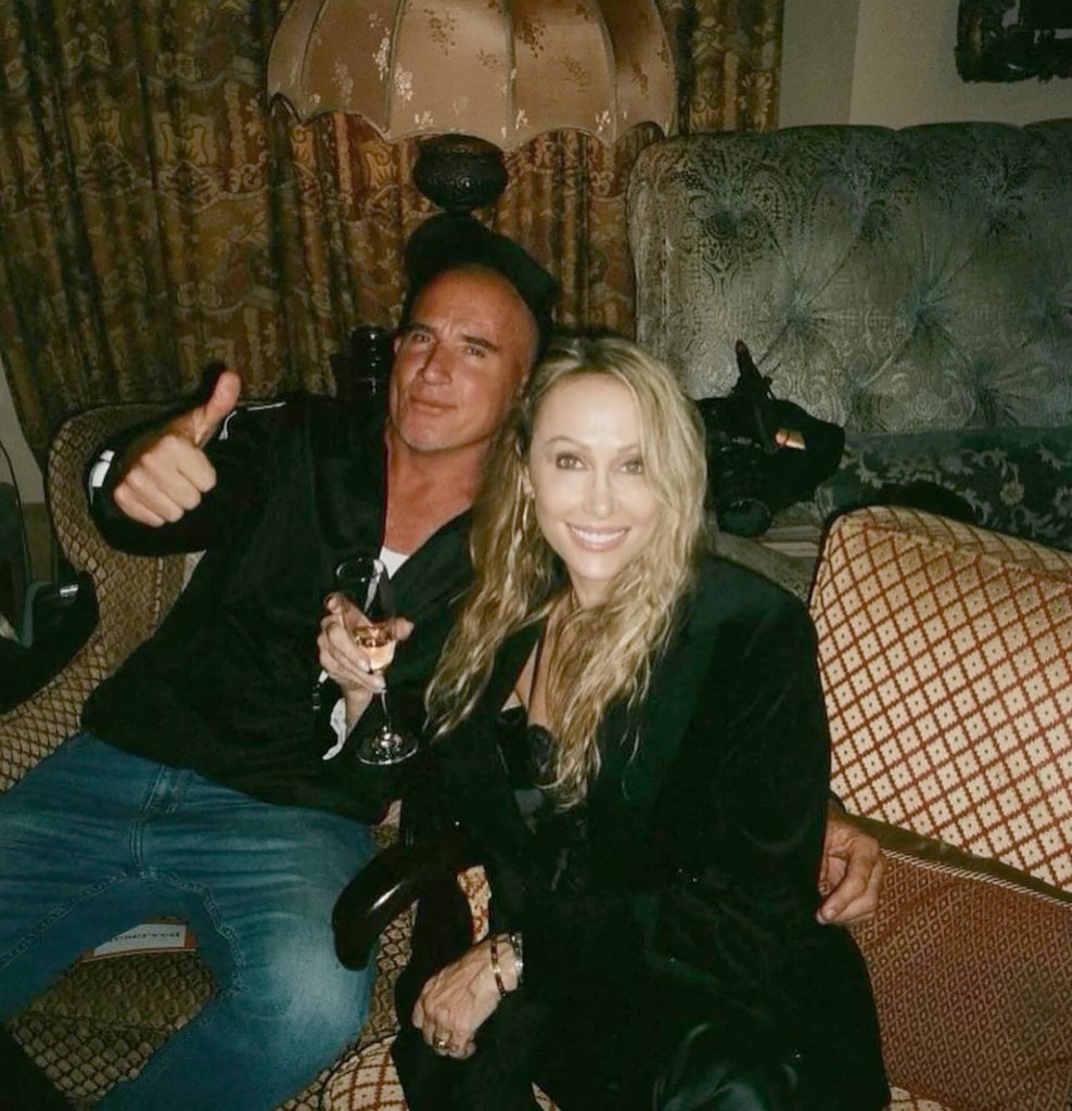Dominic Purcell and Tish Cyrus sitting together on a sofa, with Purcell giving a thumbs up.