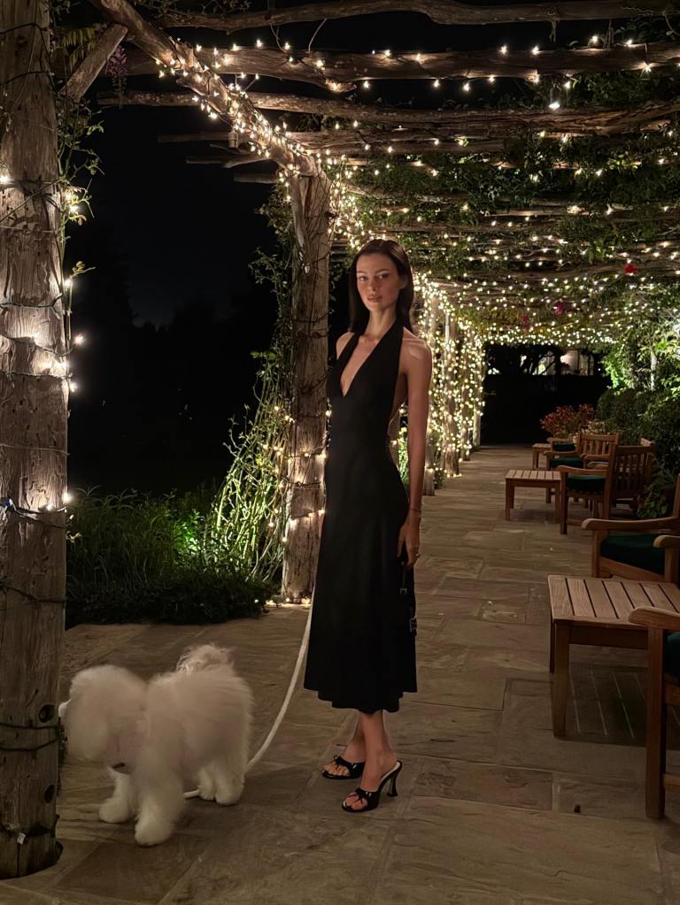 Nicola Peltz in a black dress on a patio next to a white dog under twinkling lights.