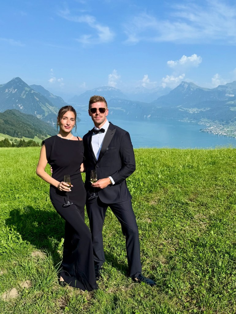 Amanda Batula and Kyle Cooke in a field with a lake and mountains in the background, holding champagne glasses.