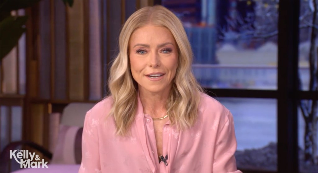 Kelly Ripa smiling while wearing a pink blouse on a TV show set.