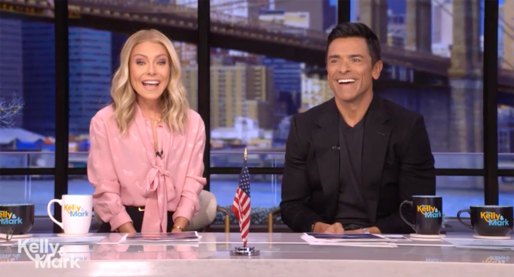 Kelly Ripa and Mark Consuelos laughing while sitting at a desk with coffee mugs.