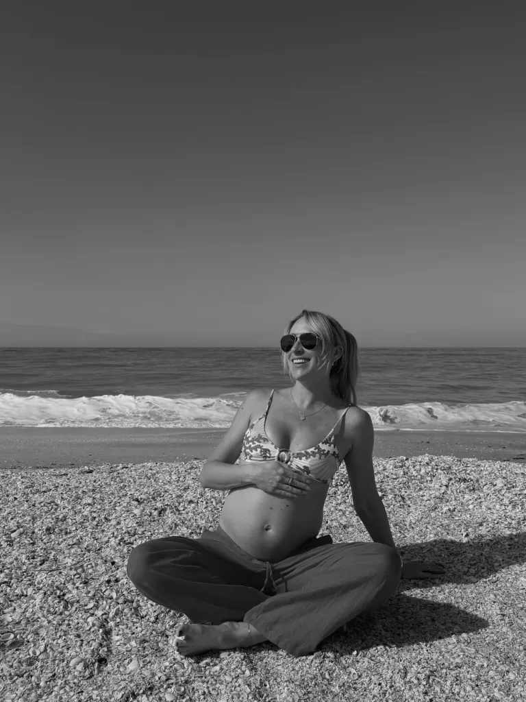 A pregnant woman in a bikini top and pants sits on a shell-covered beach, holding her baby bump and smiling at the ocean.