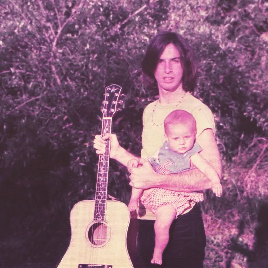 Robert Carradine holds baby Ever while balancing an acoustic guitar.