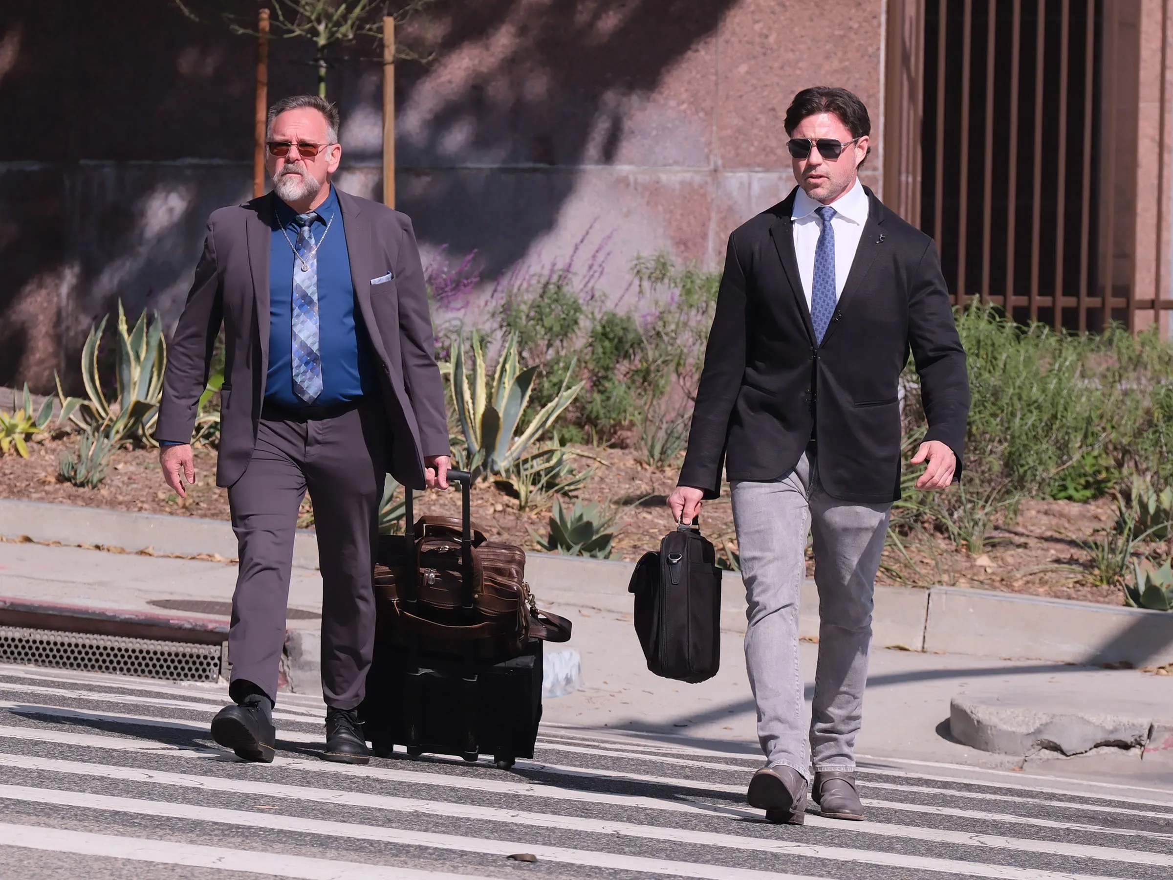 Aaron Phypers in a black suit, white shirt, and blue tie, walking with a briefcase beside another man pulling a rolling suitcase.