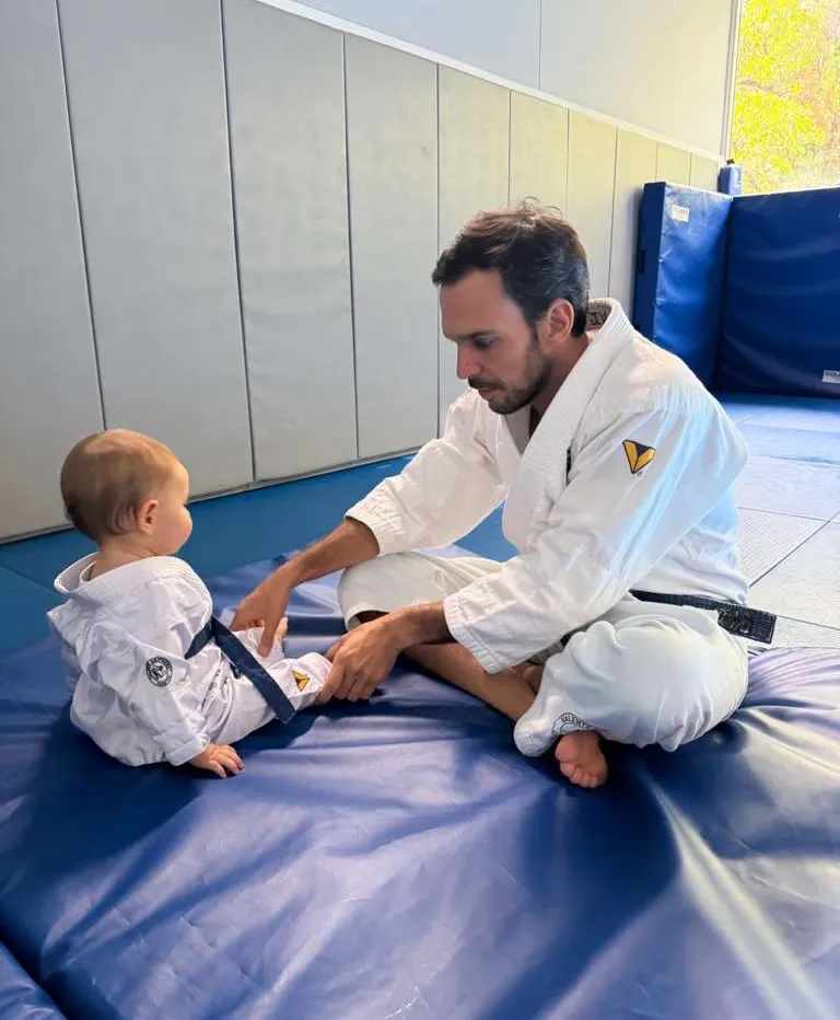 Joaquim Valente, in a black-belt gi, adjusts a baby's tiny gi while sitting on a blue mat.