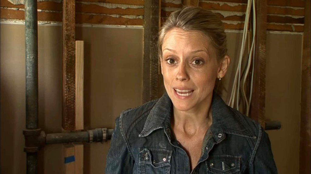 A woman wearing a denim jacket speaks in a room under construction with exposed pipes and wall studs.