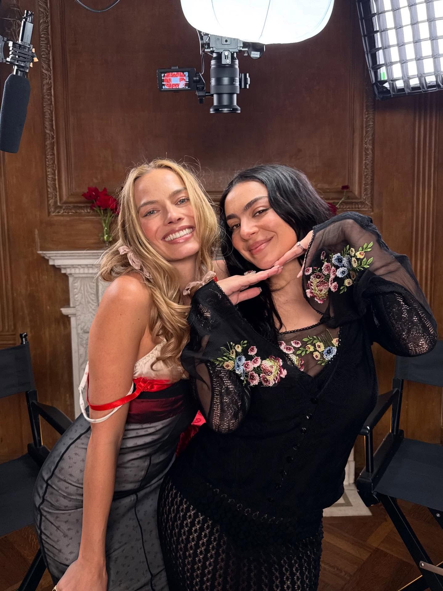 Margot Robbie and Charli XCX smiling in front of a camera and lighting equipment.