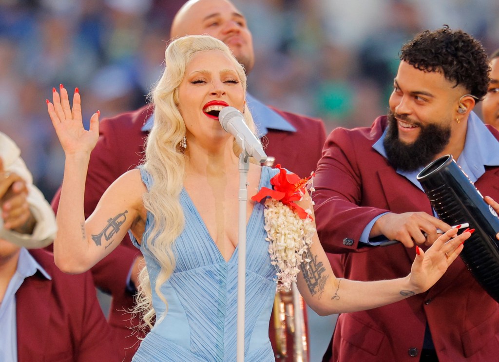 Lady Gaga singing into a microphone during a Super Bowl halftime show, wearing a light blue dress with red and white floral accents, surrounded by backup performers in maroon suits.