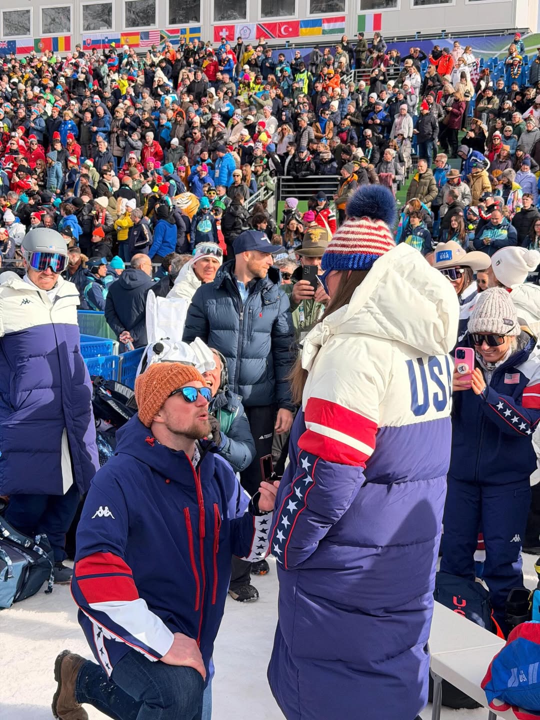 A man in a blue, red, and white jacket kneels before a woman in a matching jacket, holding up a ring in front of a stadium crowd.