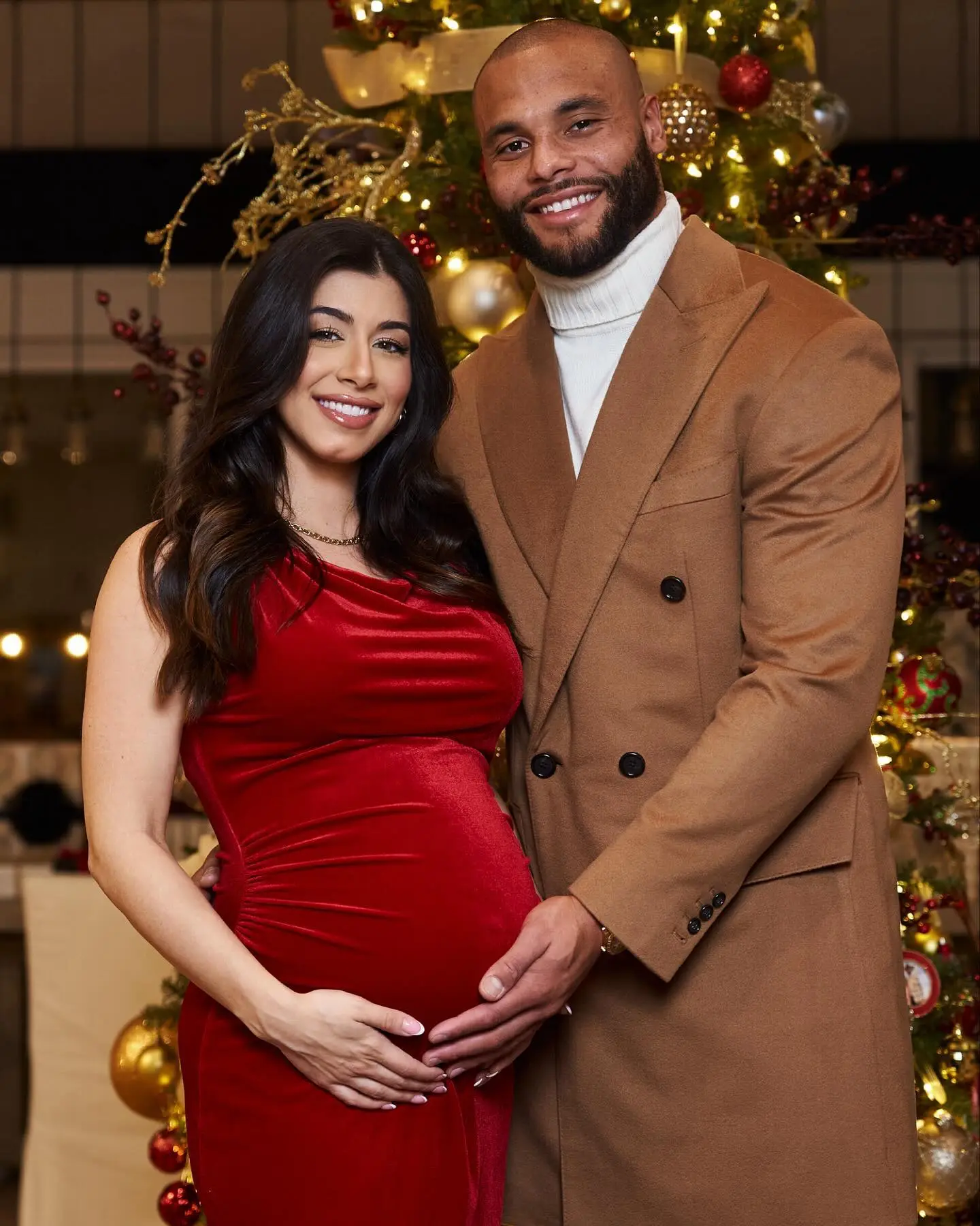 Sarah Jane Ramos and Dak Prescott posing in front of a Christmas tree, with Ramos cradling her baby bump.