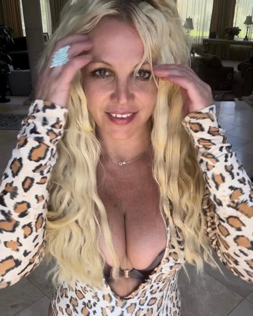 Britney Spears in a leopard print top, hands touching her blonde hair, smiling at the camera.