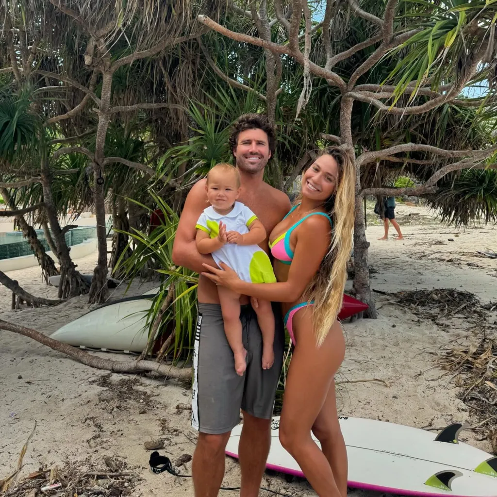 Brody Jenner, Tia Blanco, and their baby pose on a beach with a surfboard.
