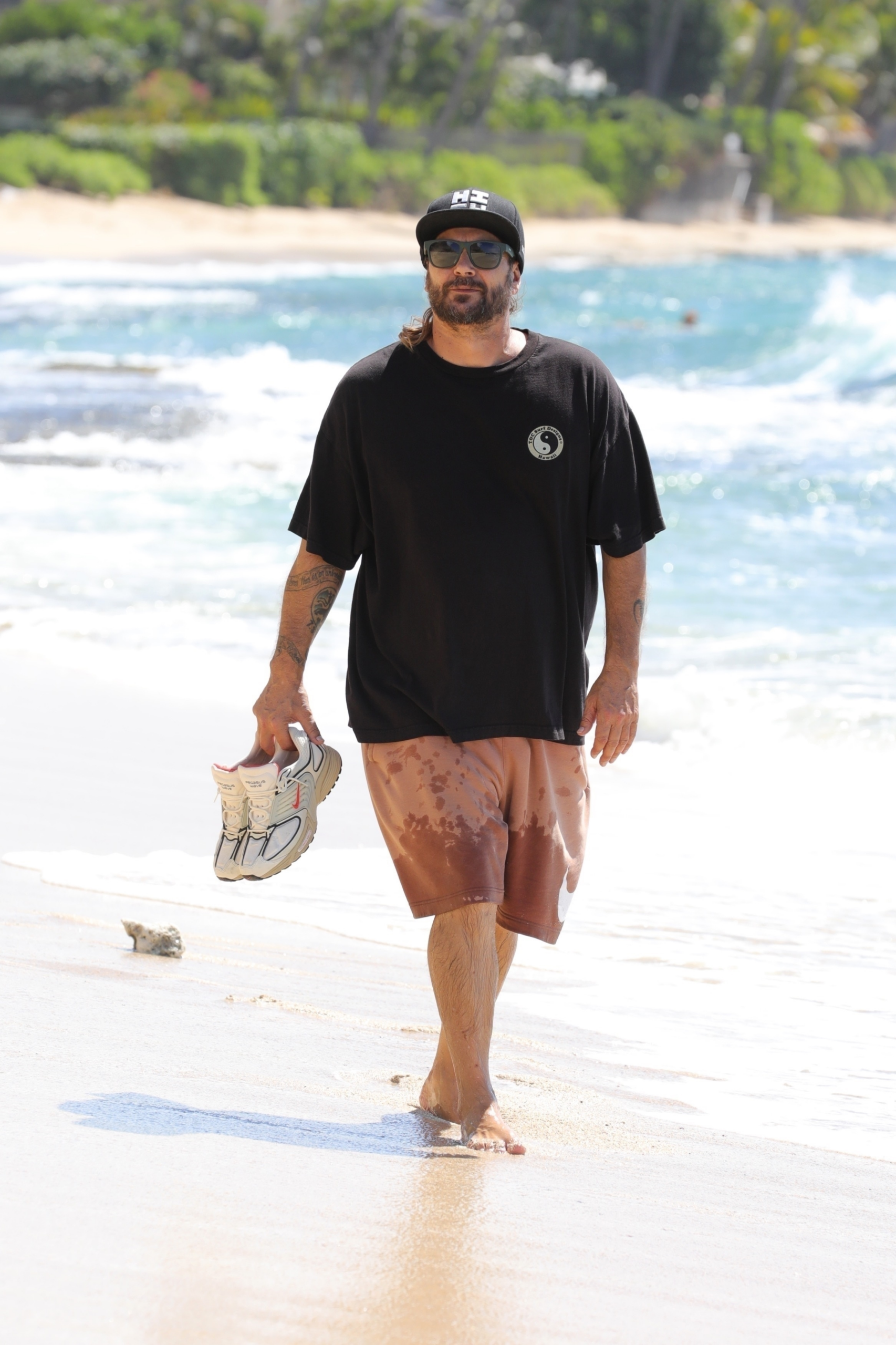 Kevin Federline walking barefoot on a beach in 2025, holding his sneakers.