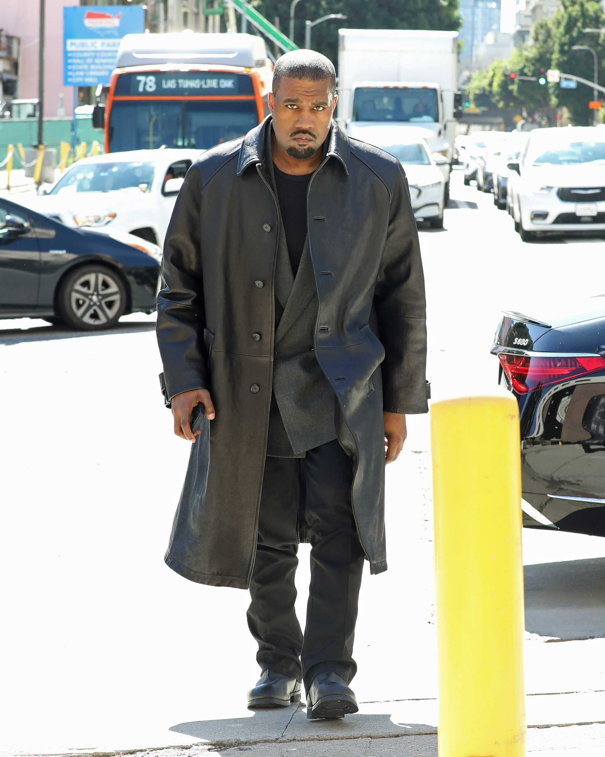 Kanye West in a long black leather coat and dark clothing with a serious expression, walking on a street in Los Angeles.