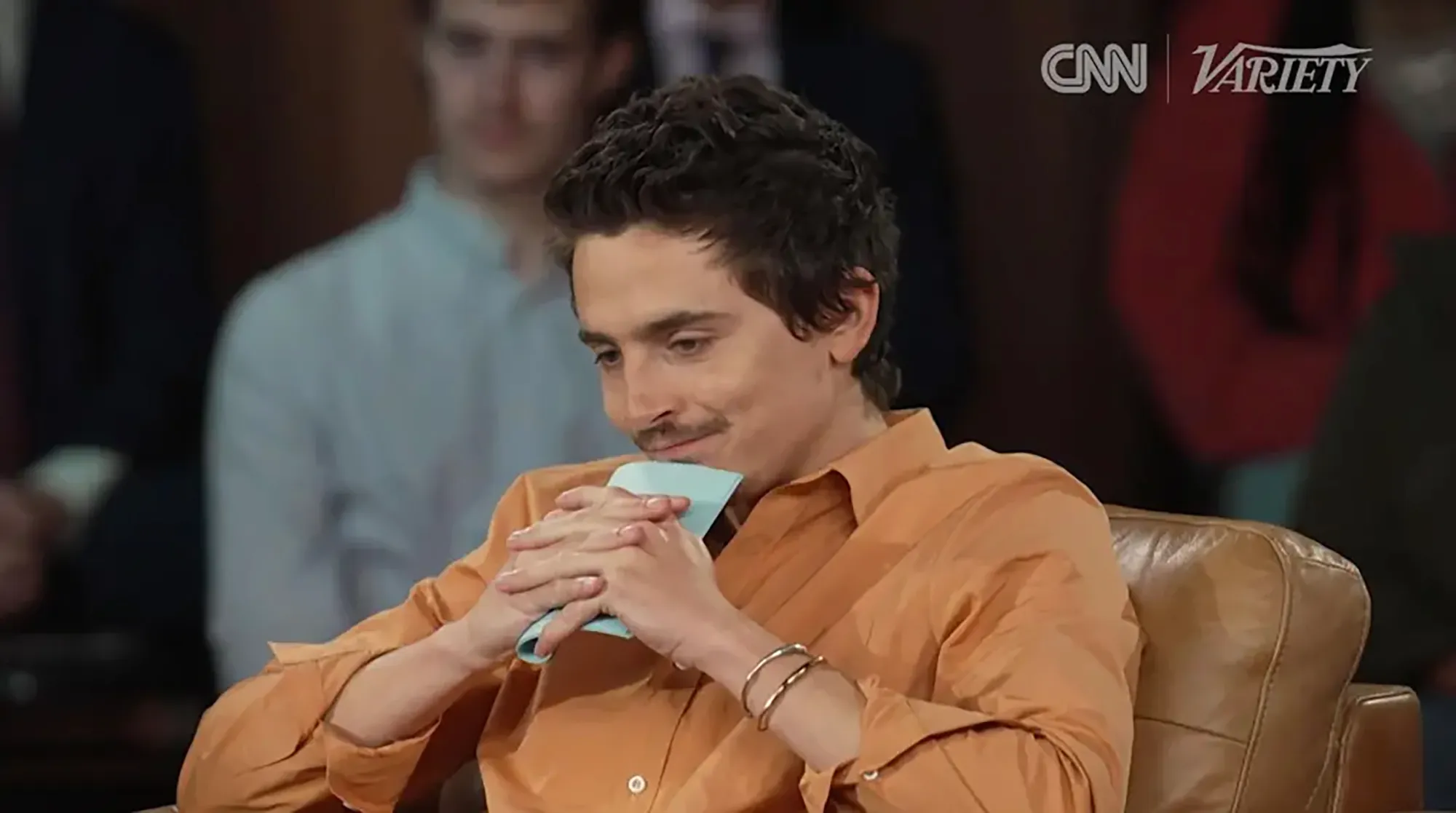Timothee Chalamet seated at a CNN town hall, wearing an orange shirt and looking down.