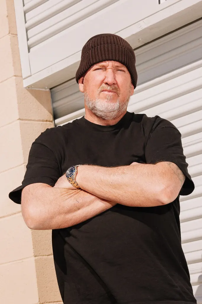 Darrell Sheets wearing a brown beanie and black shirt, arms crossed in front of a storage unit door.