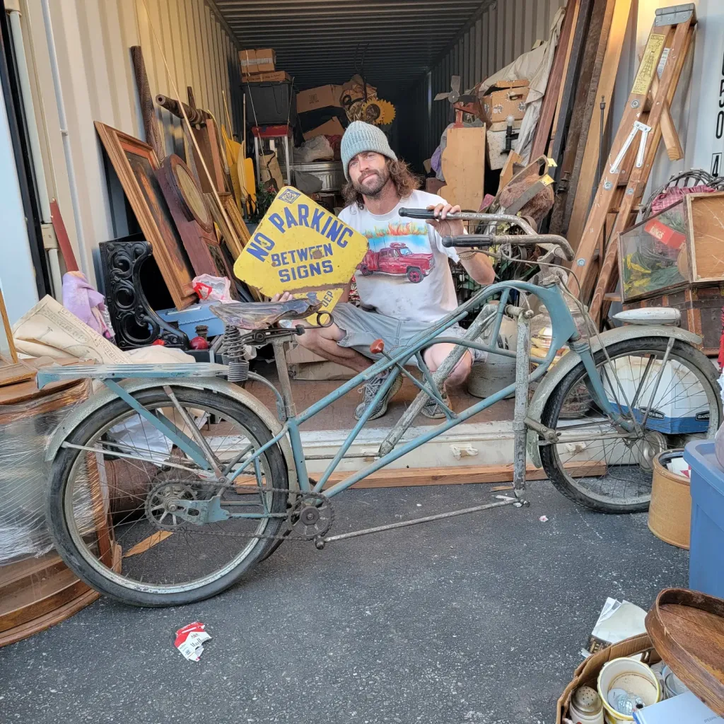 Dusty Riach holding a NO PARKING BETWEEN SIGNS sign in a storage unit beside a vintage bicycle.