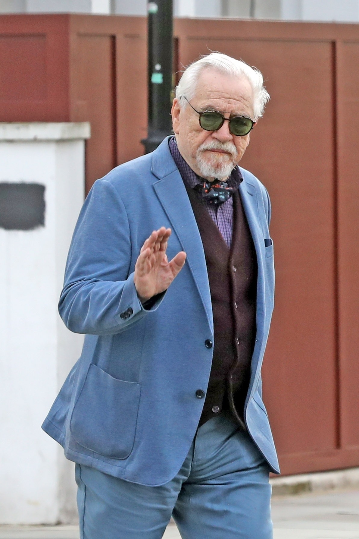 Brian Cox in London wearing a blue blazer, maroon vest, and patterned scarf, waving to the camera.