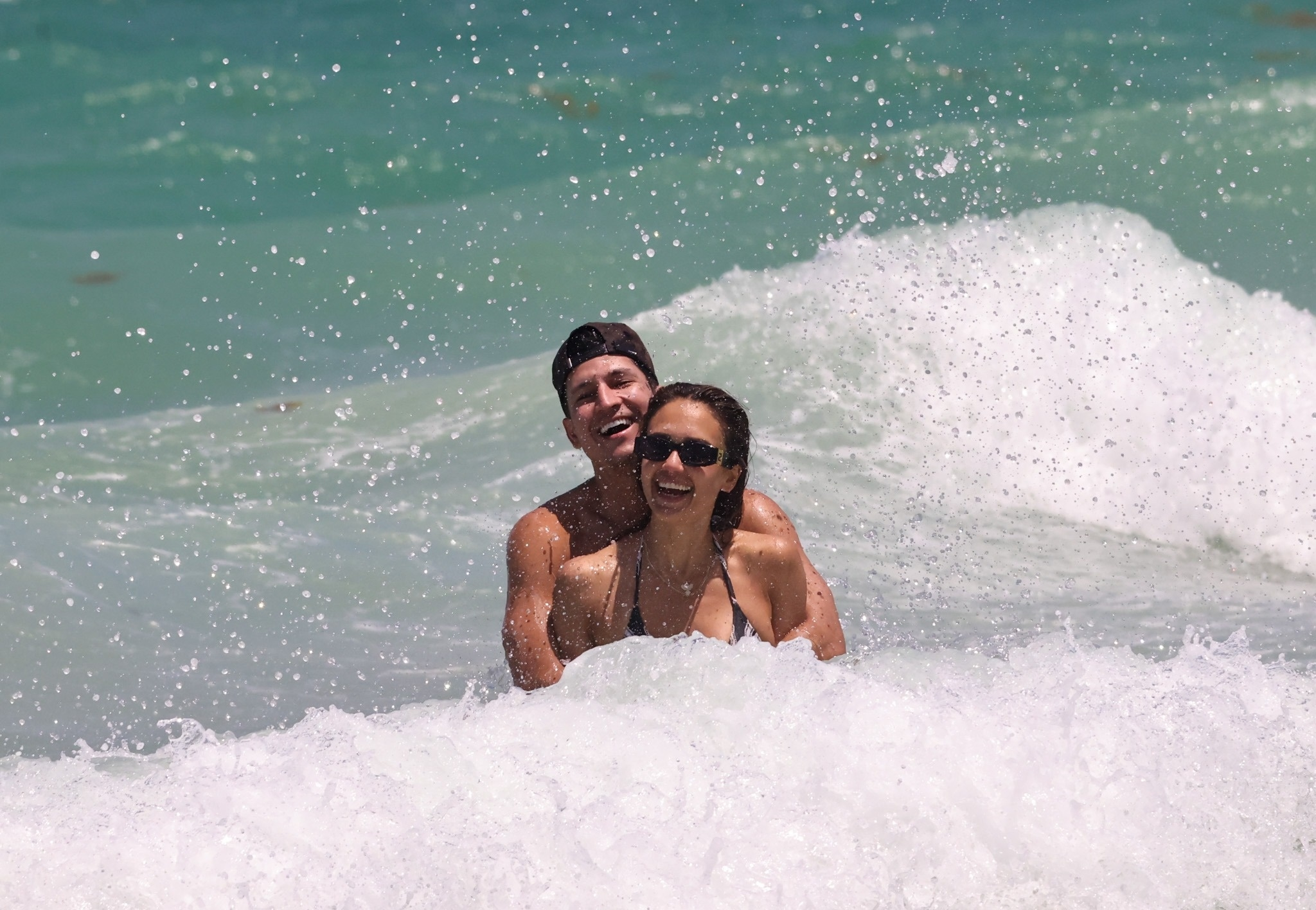 Jessica Alba and Danny Ramirez laughing in the ocean with waves breaking around them.