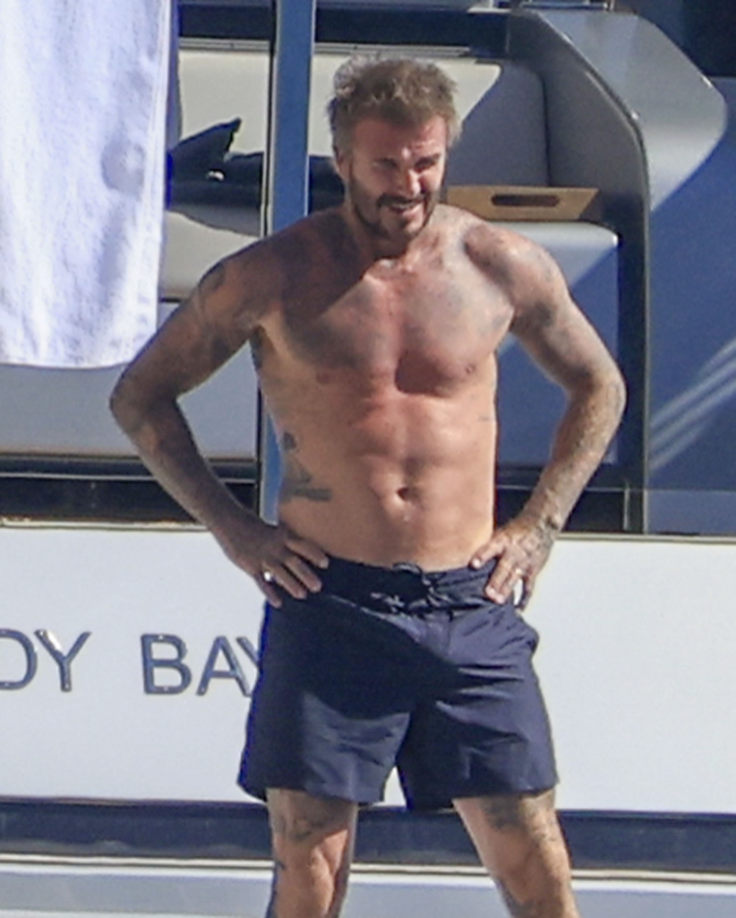 David Beckham stands shirtless on the yacht in navy swim trunks.