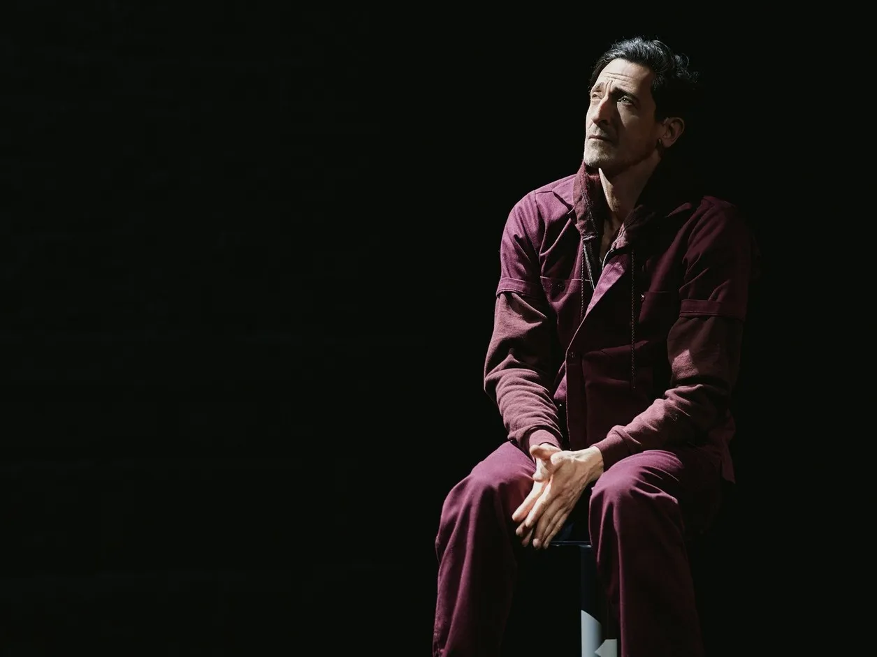 Adrien Brody in a maroon jumpsuit, sitting on a chair, looking up.