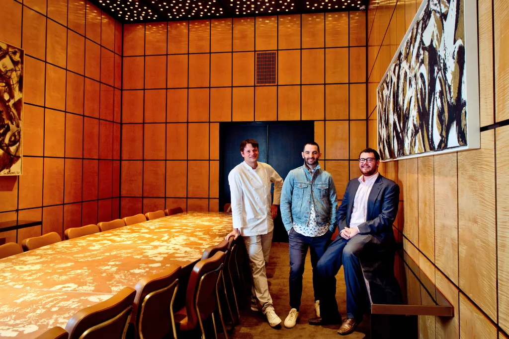 Three men, including a chef, in a wood-paneled room with a long table.