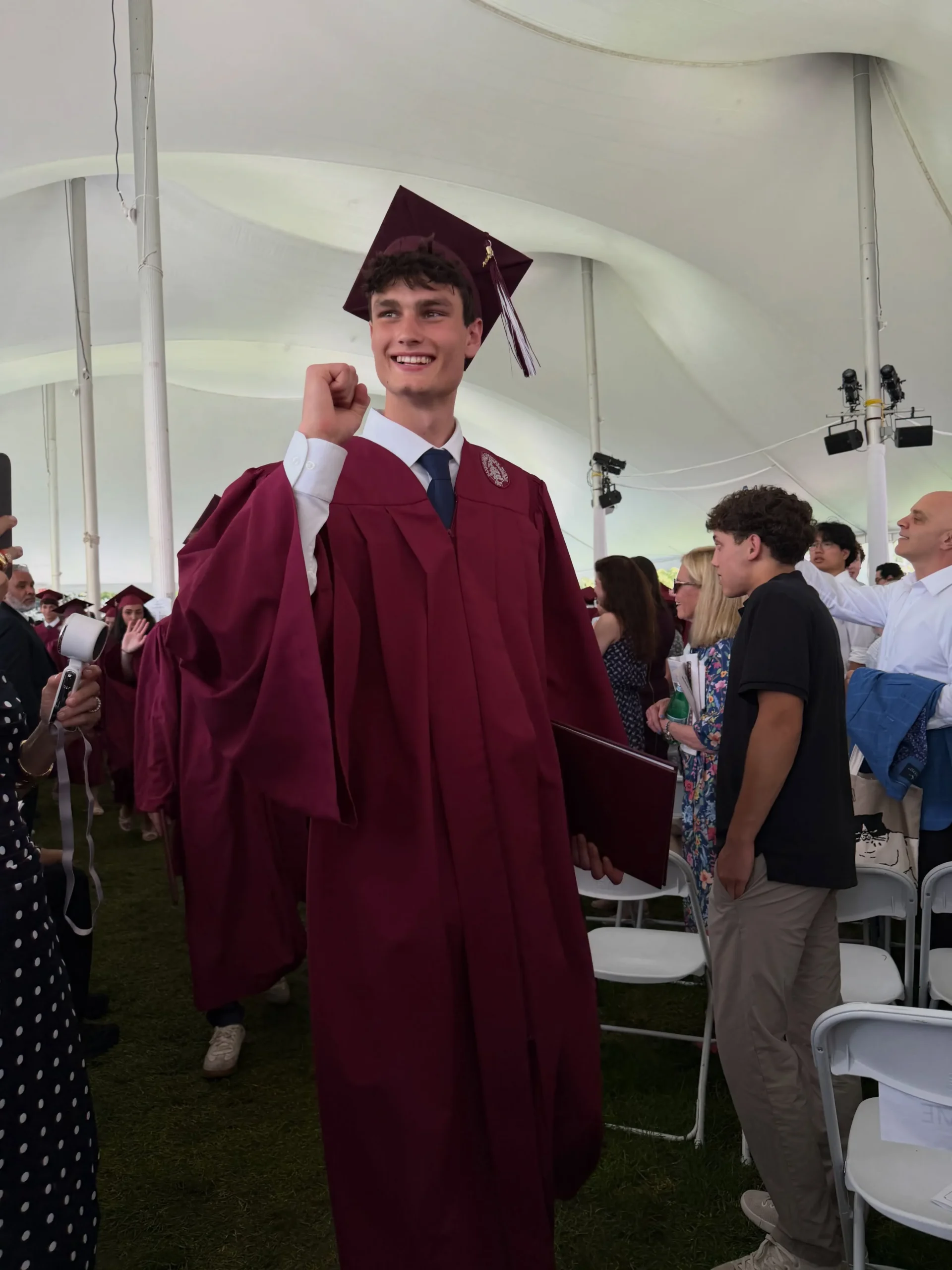 Jack Moynahan at his graduation ceremony, wearing a maroon gown and cap, smiling and holding a fist up.