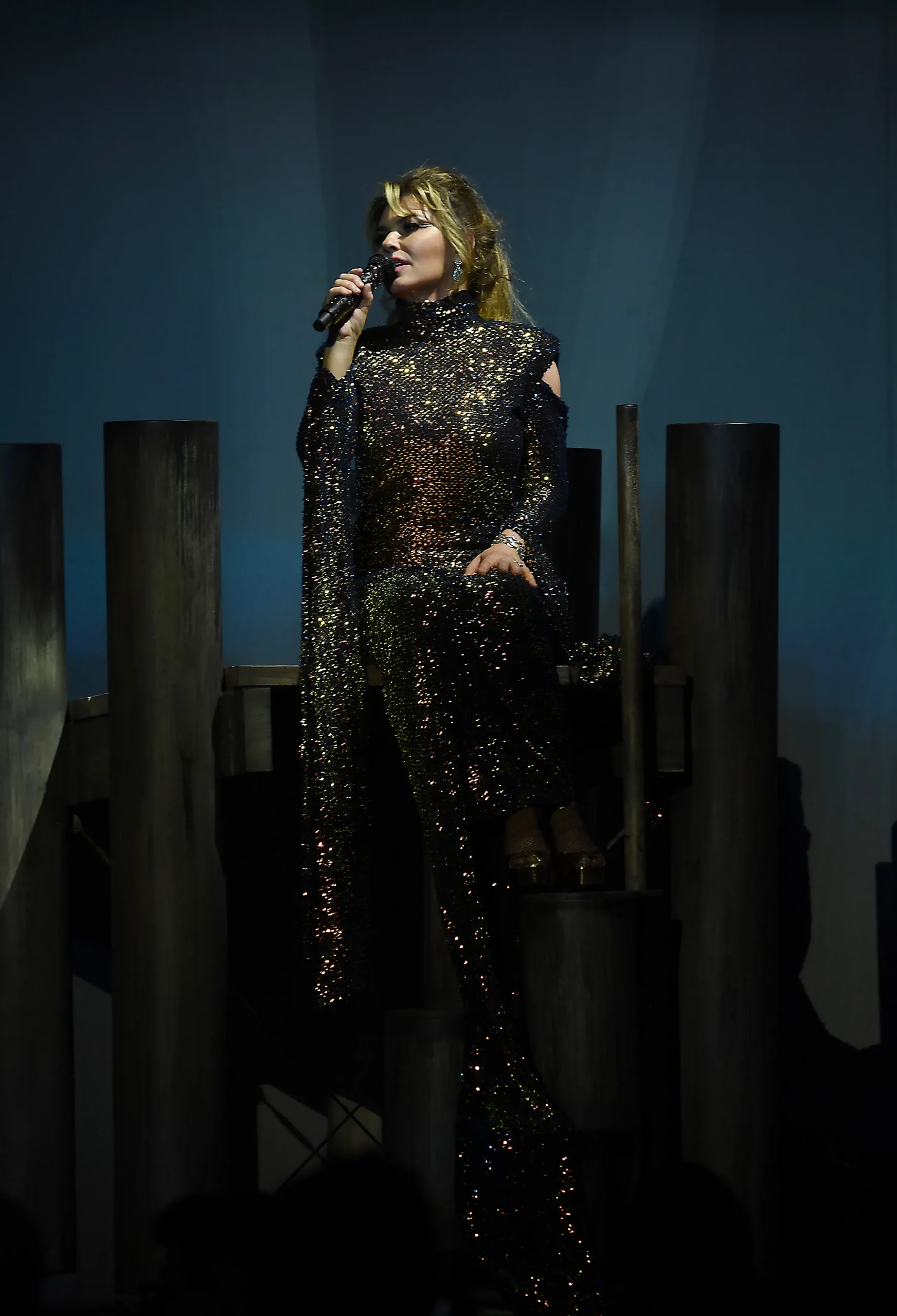 Singer Shania Twain performs in a long black sequined gown and holds a microphone.