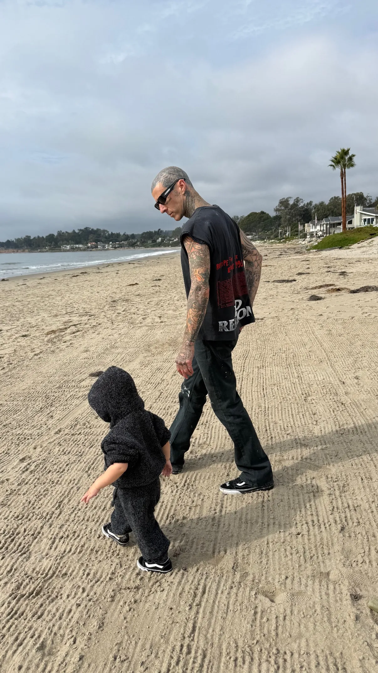 Travis Barker walking on a beach with his child.