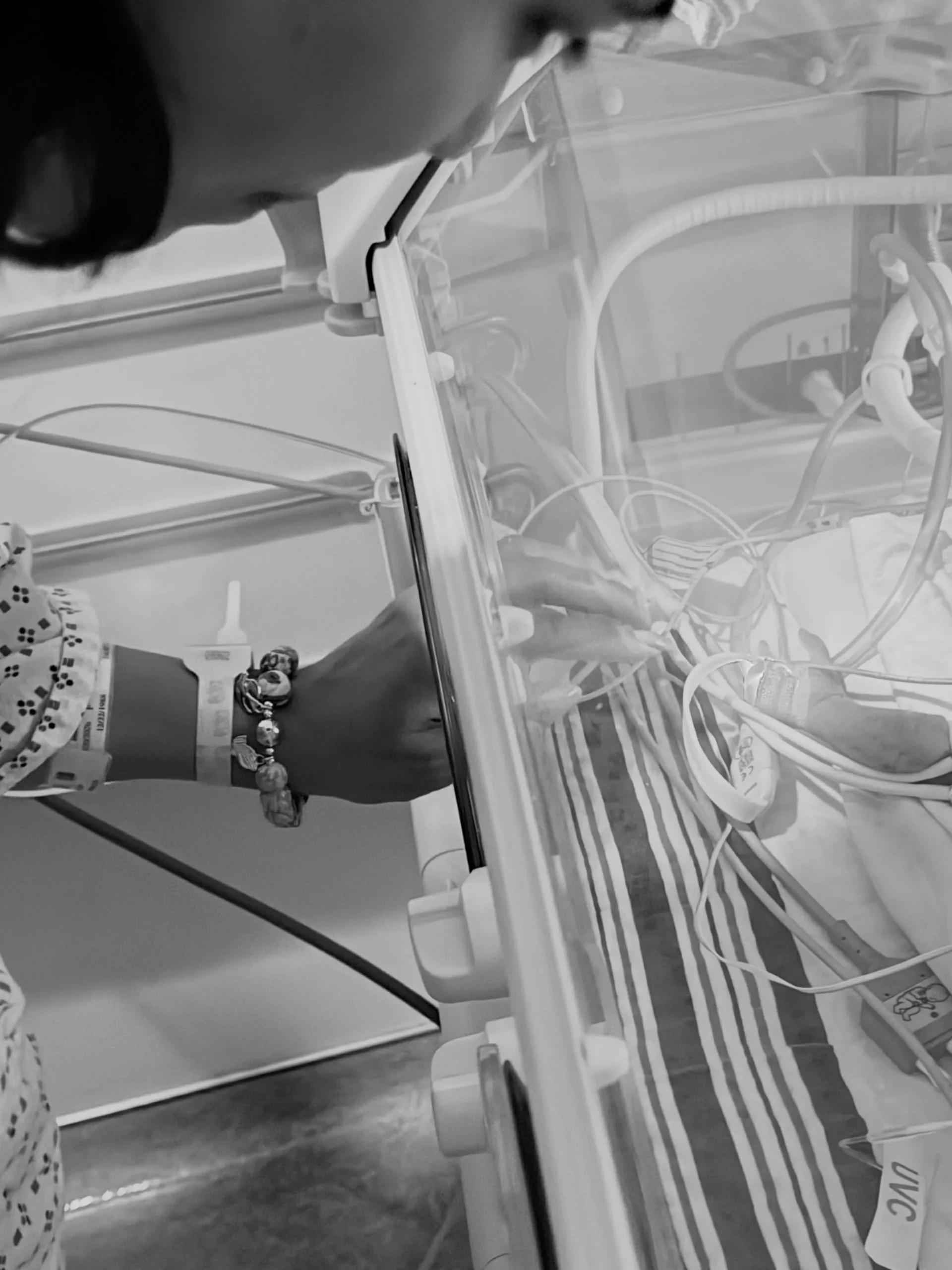 A parent's hand with a hospital bracelet reaching into a NICU incubator to touch their premature baby.