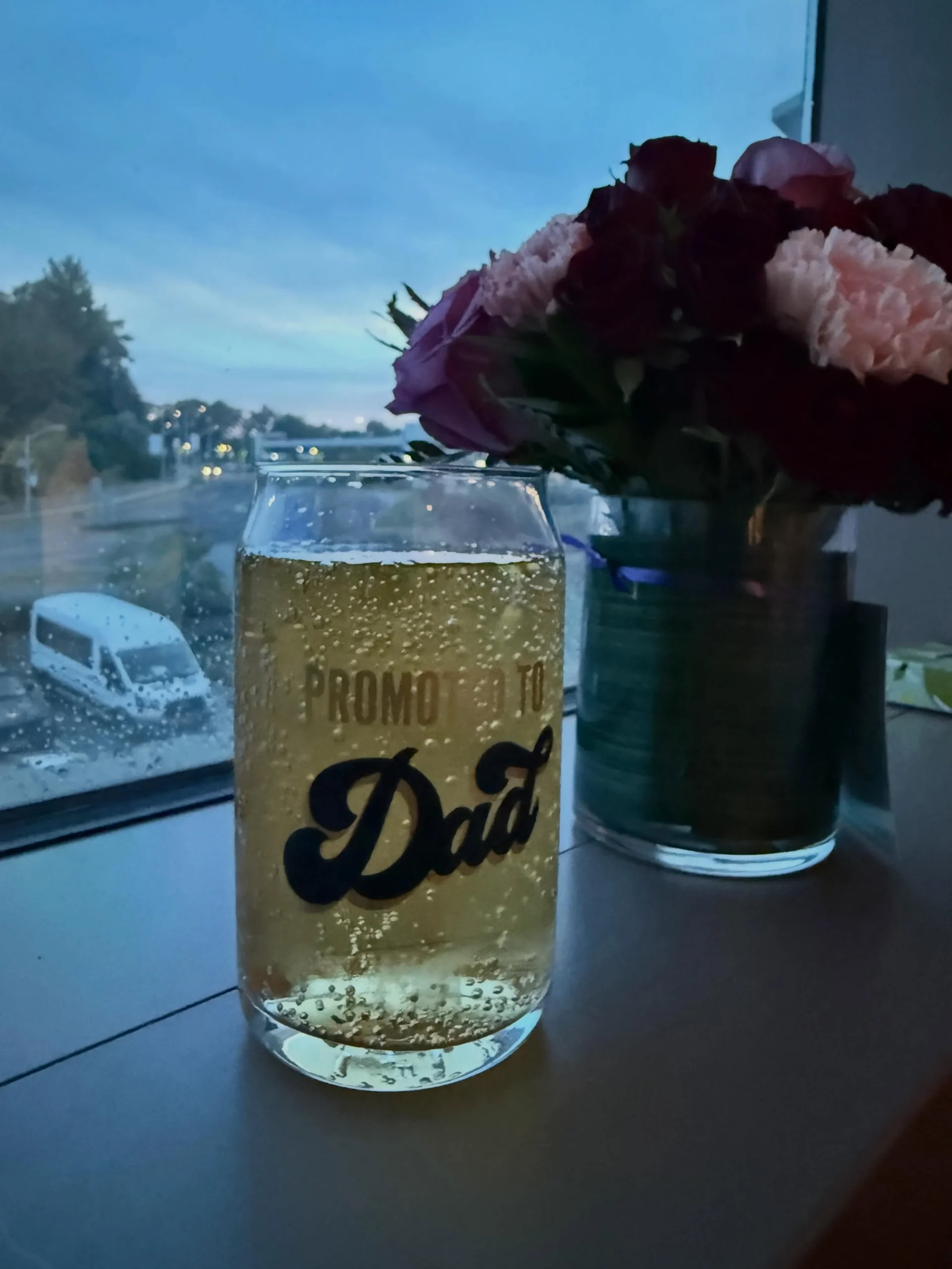 A celebratory glass reading "Promoted to Dad" beside flowers, shared as part of the couple's birth announcement.